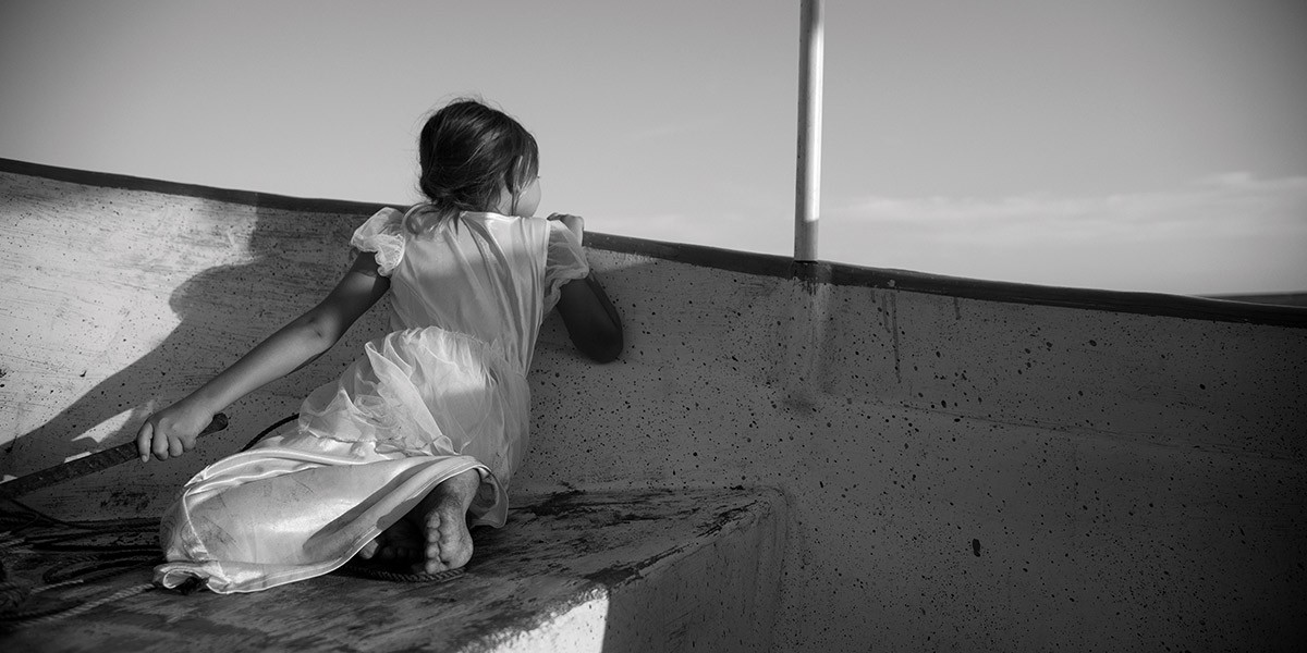 Young girl in a white dress sits barefoot on a boat, looking over its side at the water.