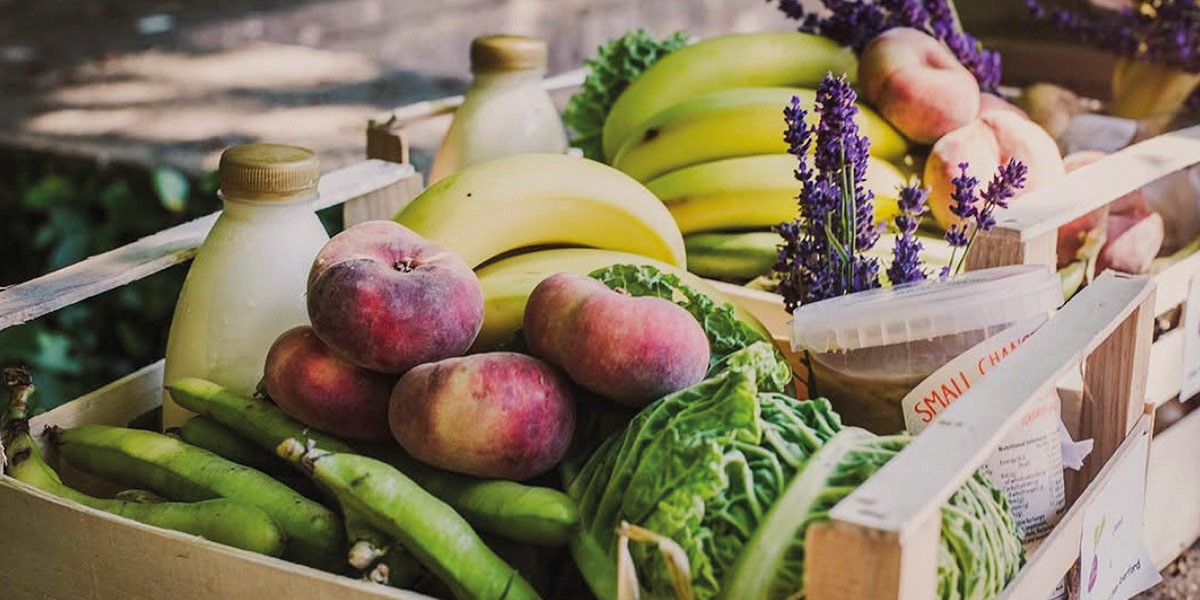 Wooden crate filled with peaches, bananas, green beans, lettuce, and purple flowers outdoors.