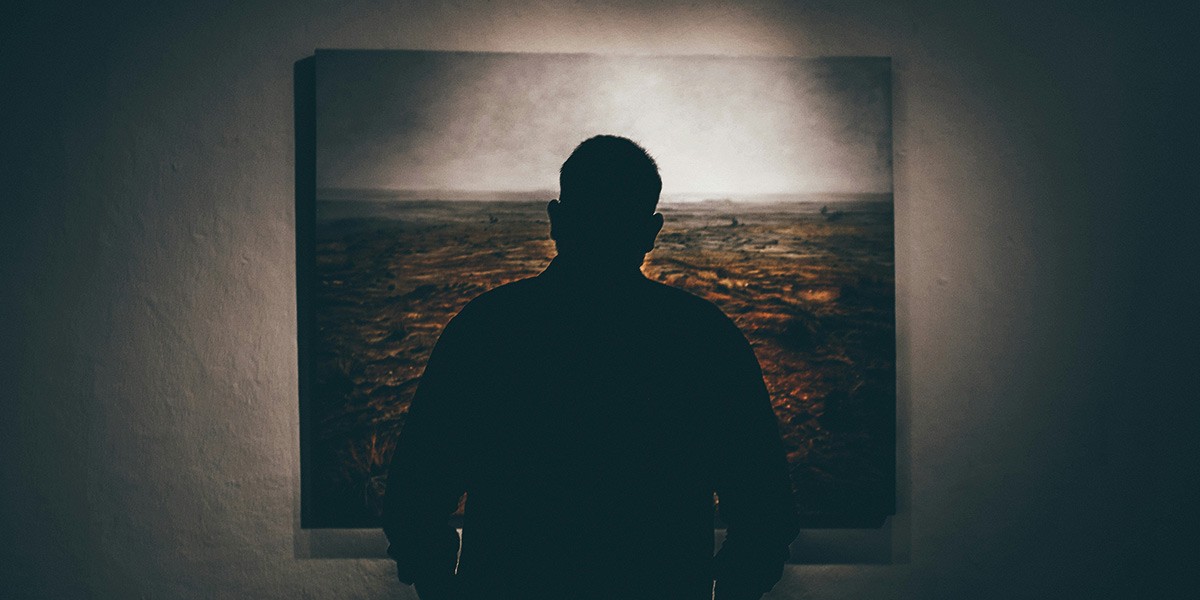 Silhouette of a person standing and looking at a large framed landscape artwork on a wall.
