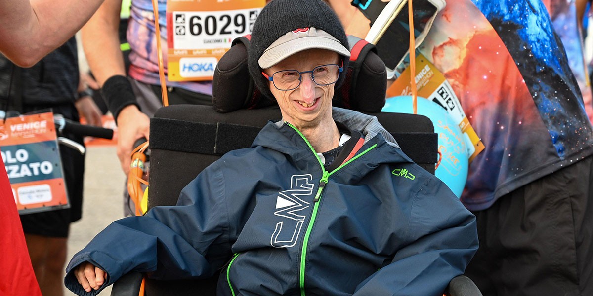 Smiling man in a wheelchair wearing a navy jacket and cap, surrounded by marathon participants.