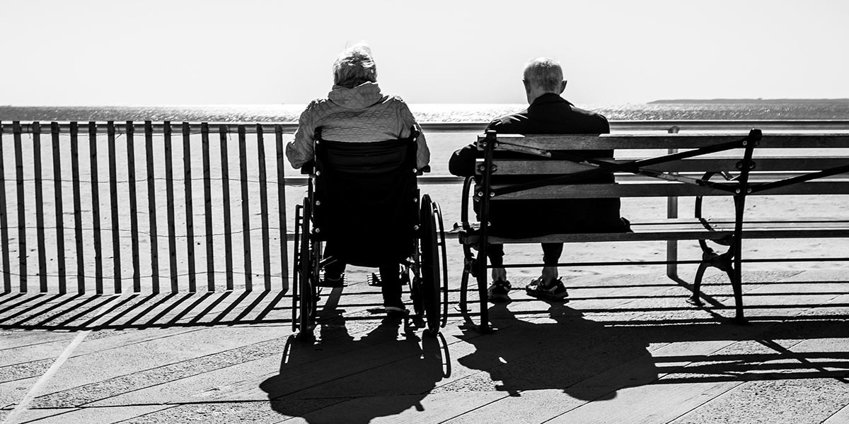 Black and white photo of two elderly people facing a beach, one in wheelchair, one on bench.