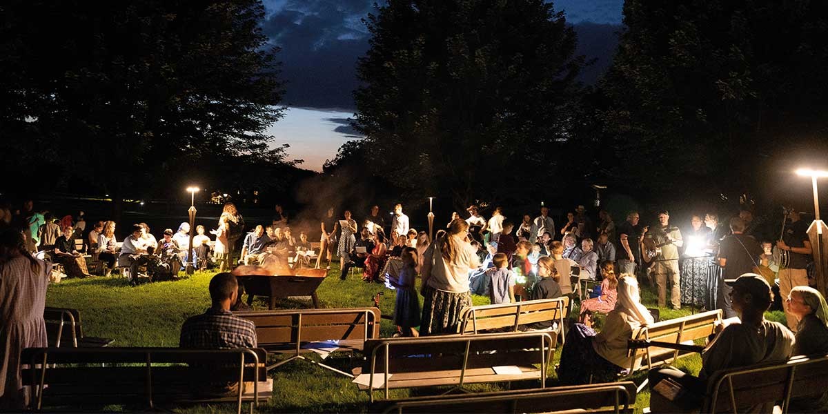 Large group of people gathered on benches around a fire pit outdoors at dusk with lamps illuminating them.