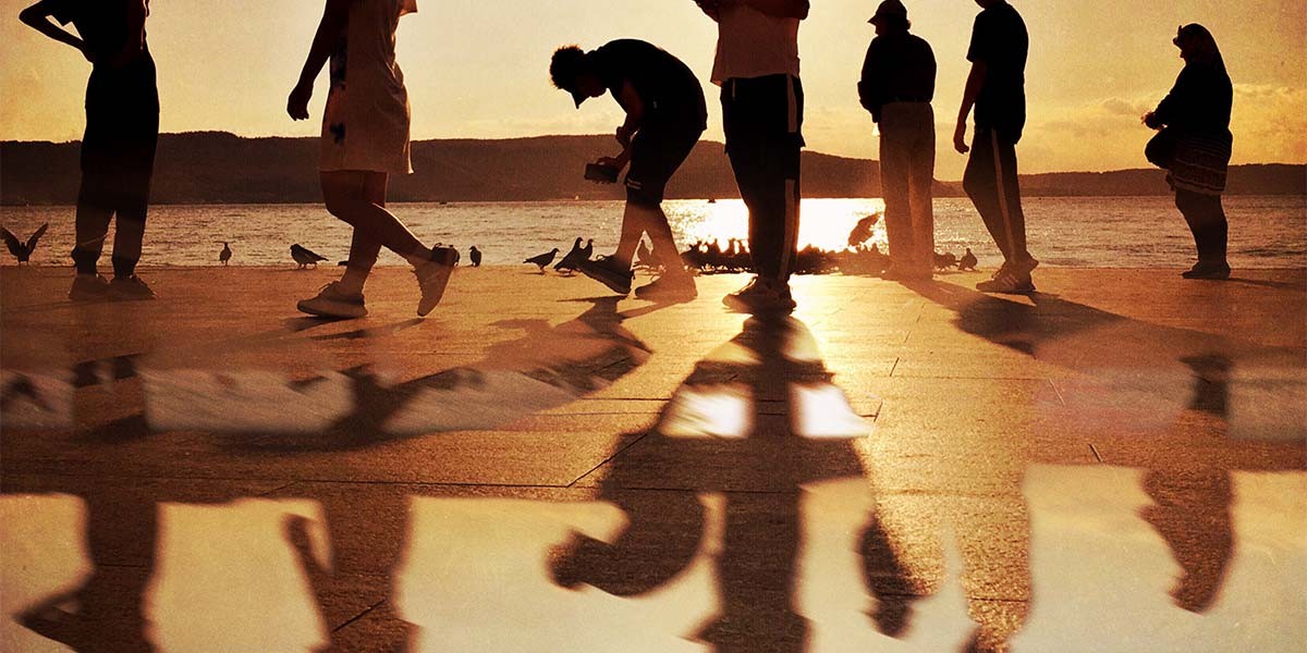 Silhouettes of people walking and standing by the water at sunset with their elongated shadows on the ground.