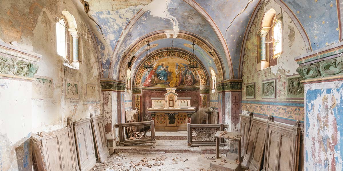 Interior of an abandoned ornate chapel with peeling paint, wooden pews, and religious murals.