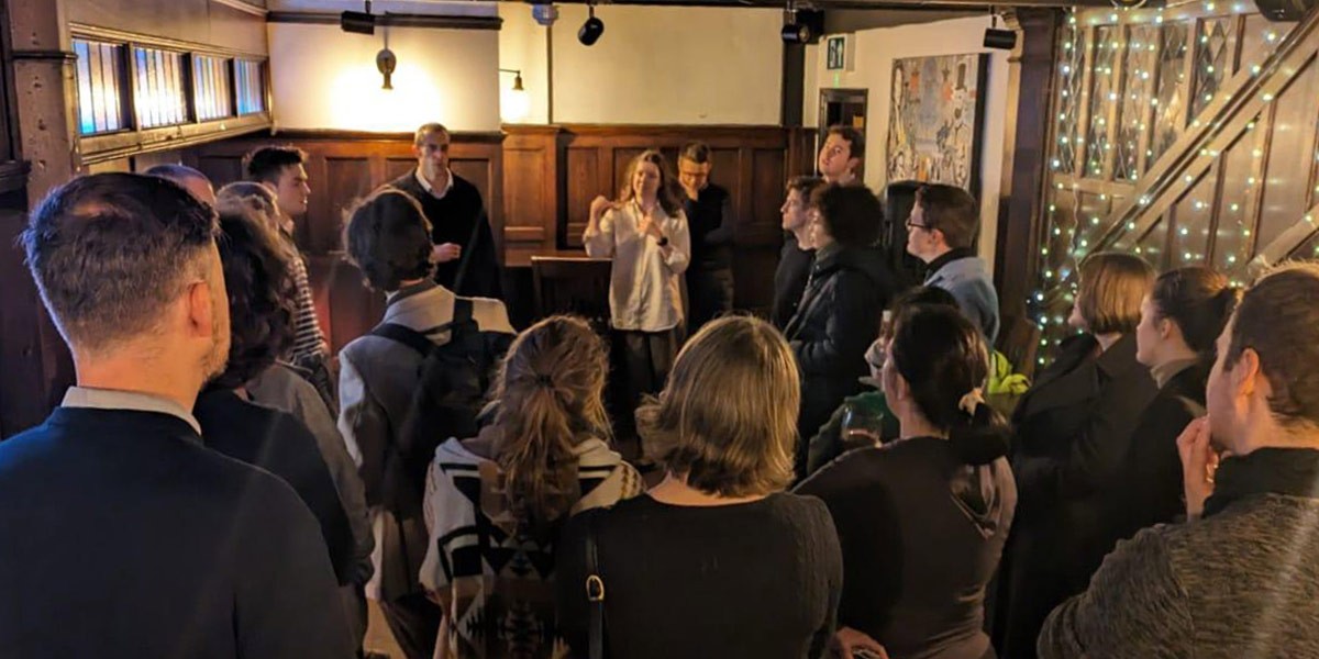 Group of people gathered indoors, attentively listening to a woman speaking in front of them.
