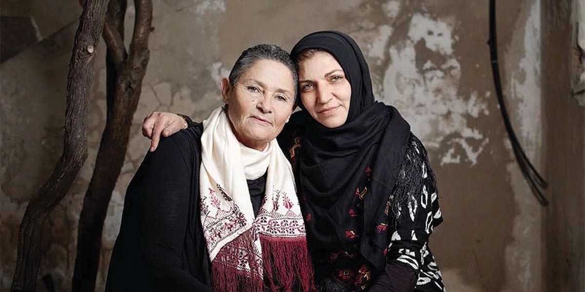 Two women sitting close with their arms around each other, one wearing a black hijab and the other a white scarf.