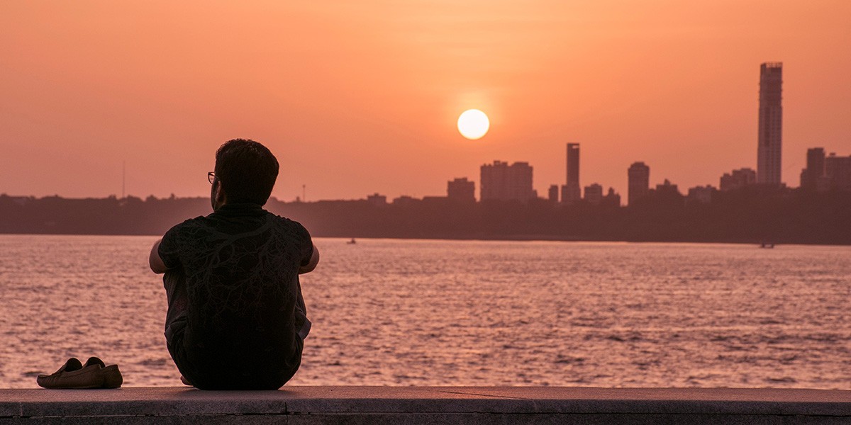 Man sitting by water's edge, watching sunset over city skyline with shoes beside him.