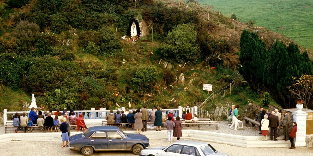 People gathered outdoors in front of a grotto with a statue of the Virgin Mary in a grassy hillside.