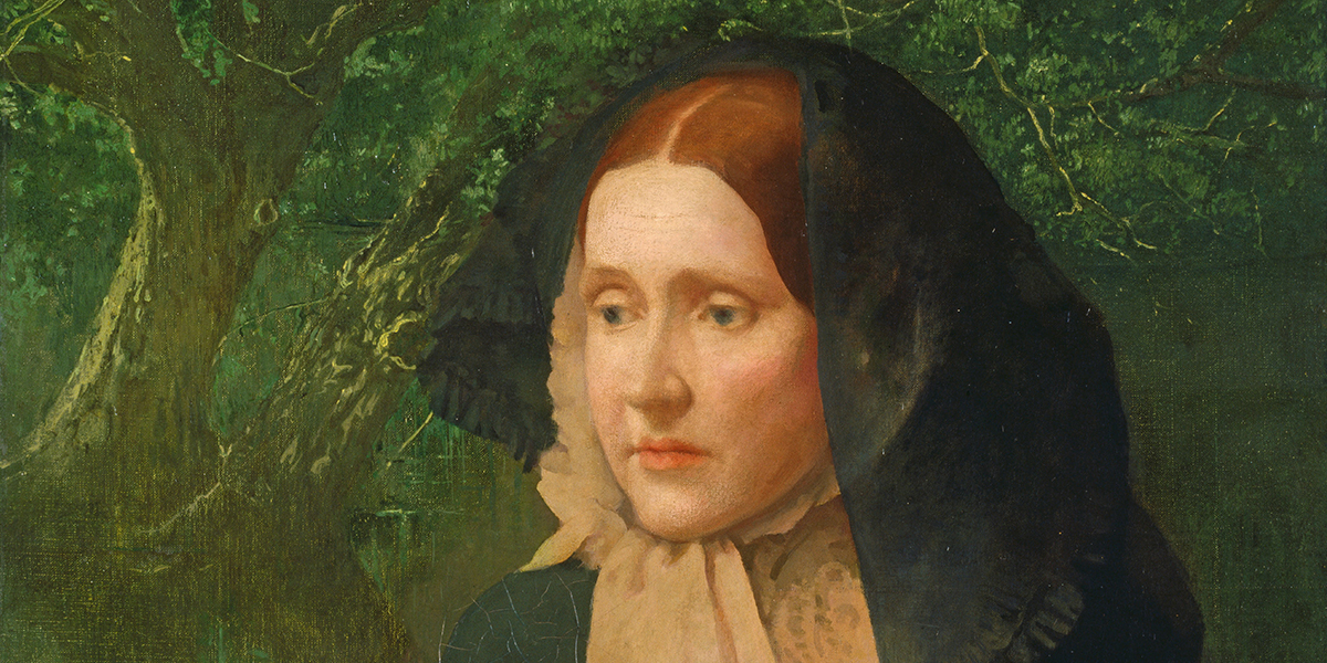 Portrait of a woman with red hair wearing a black hood and a beige scarf, standing against leafy trees.