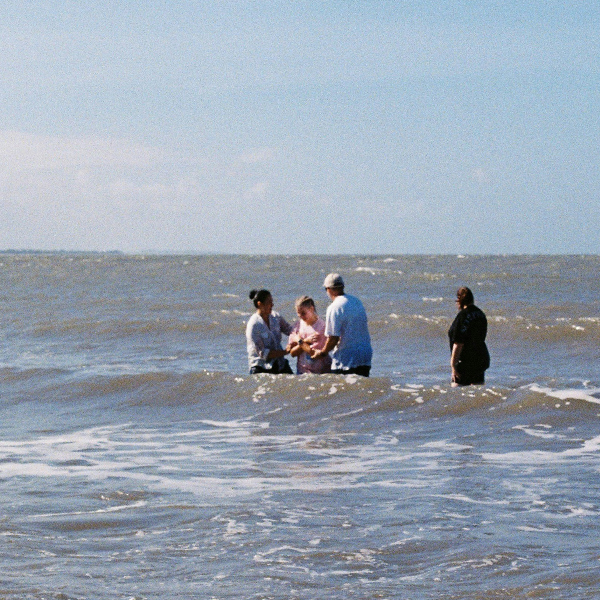 Four people standing in waist-deep ocean water on a sunny day, appearing to have a conversation.