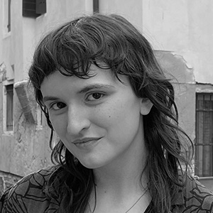 Black and white portrait of a person with wavy hair smiling slightly in an urban outdoor setting.
