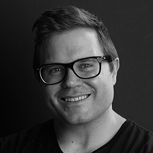 Smiling man wearing rectangular glasses and a black shirt against a dark background.