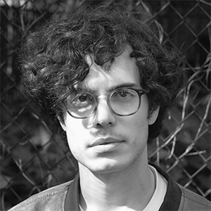 Young person with curly hair and glasses looking at the camera in front of a chain-link fence.