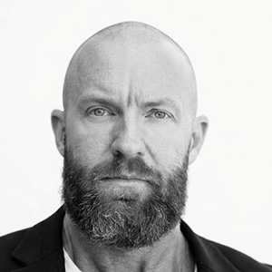 Close-up grayscale photo of a bald man with a full beard wearing a dark jacket and white shirt.