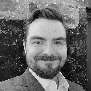 Smiling man with beard and mustache wearing a suit jacket and white shirt in front of a stone wall.