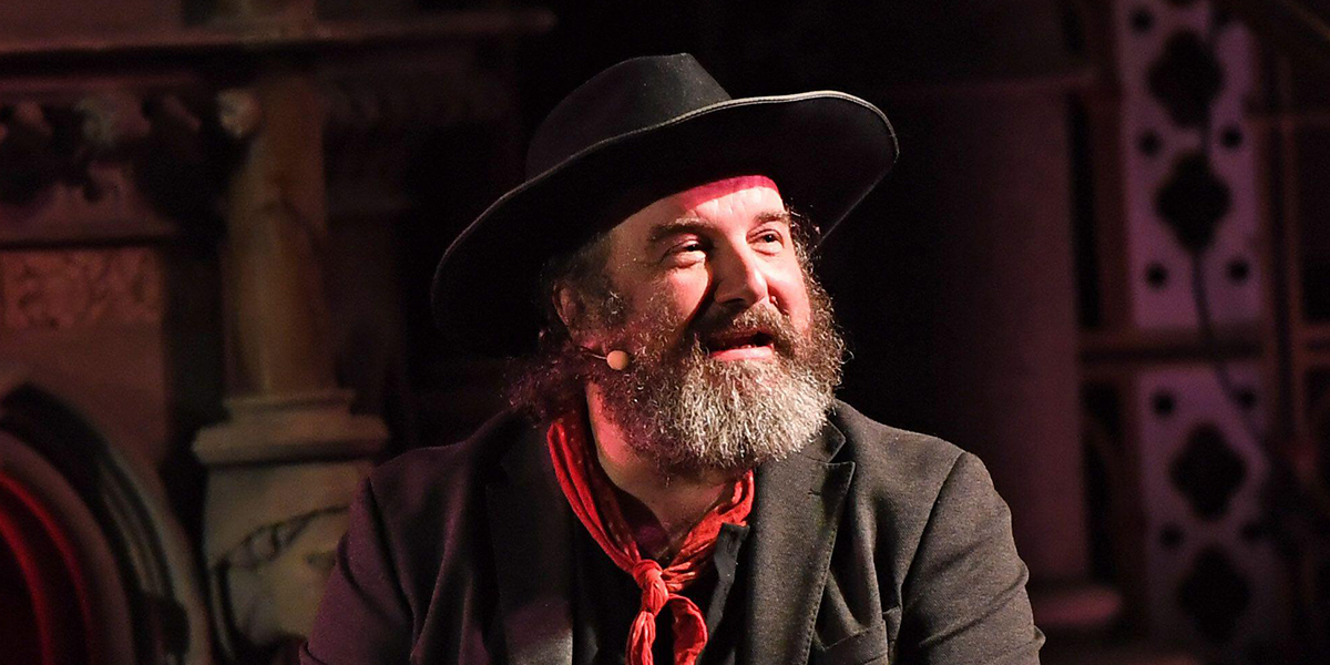 Bearded man wearing a black hat and red neckerchief speaking with a microphone on stage.