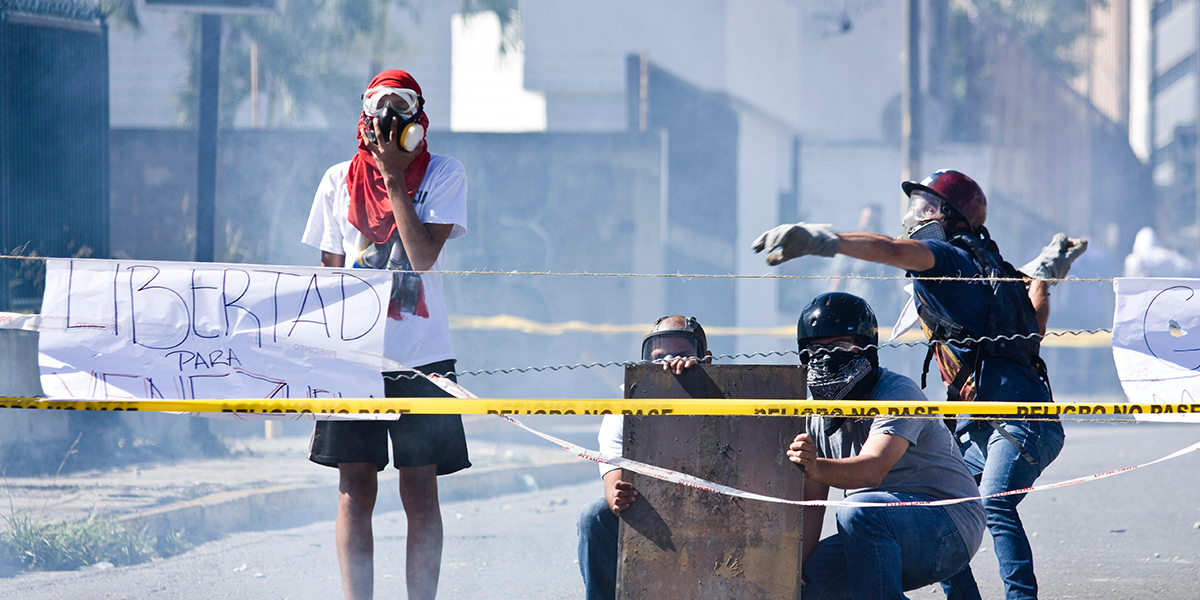 Protesters wearing masks and helmets behind barricades and caution tape, one holds a sign reading 'LIBERTAD'.