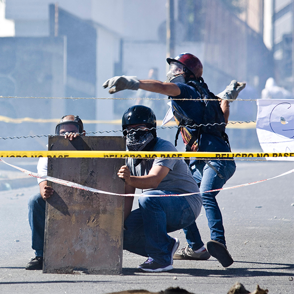 Three masked protesters behind makeshift shield and caution tape during a street confrontation.