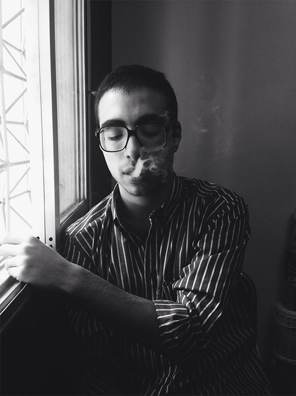Man wearing glasses, eyes closed, smoking near a window with striped shirt in black and white photo.