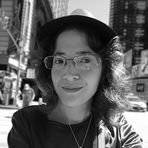 Smiling person wearing glasses and a hat in an urban setting with buildings and cars behind.