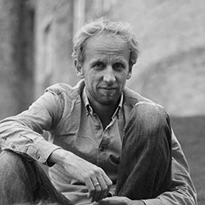 Black and white photo of a man sitting casually with one knee up, wearing a buttoned shirt and jeans.