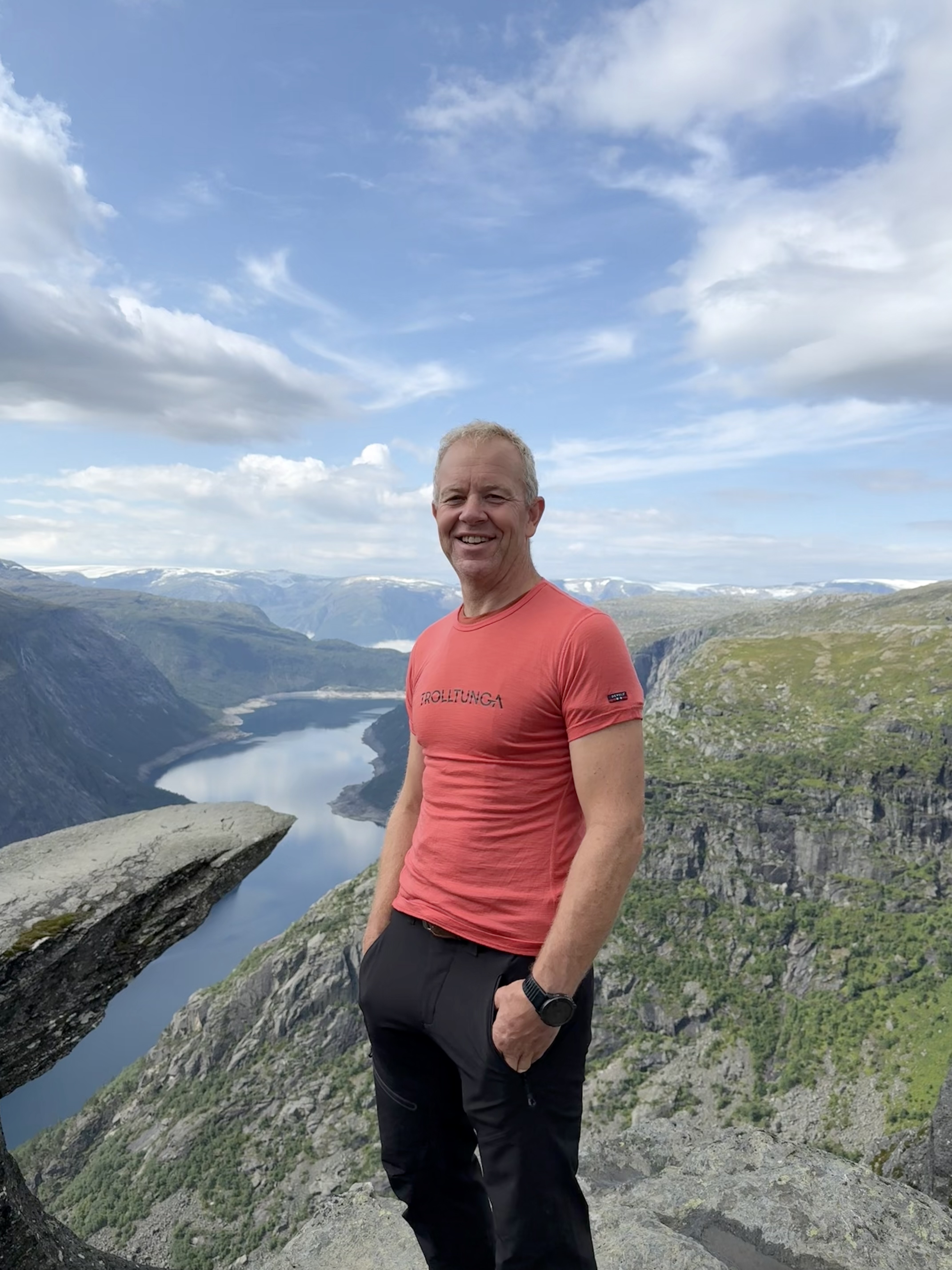 Mann i rød T-skjorte står på fjell utenfor Trolltunga med fjord og fjell i bakgrunnen.