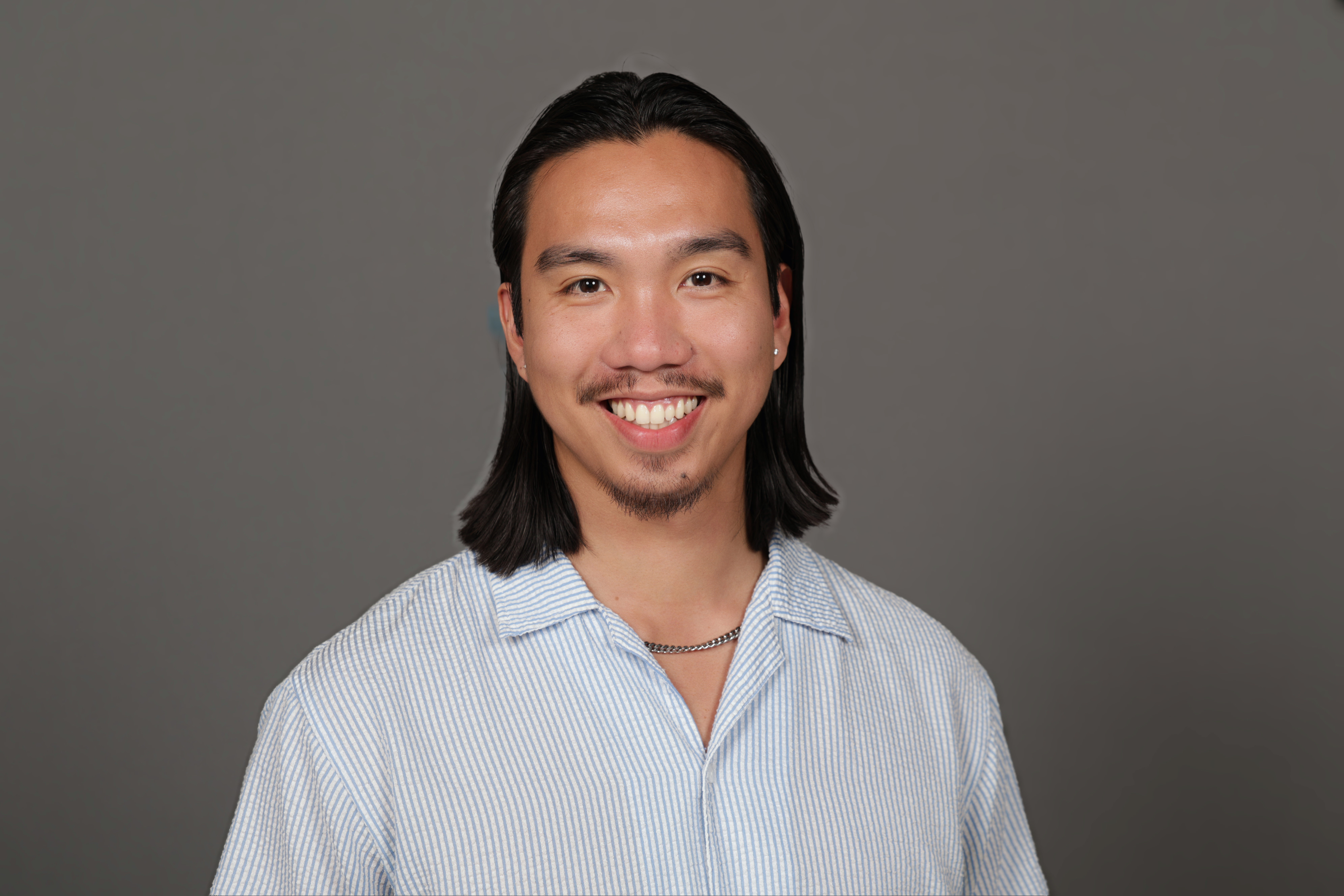 Picture of June Castro - a smiling man with medium-length black hair and a striped shirt against a gray background.