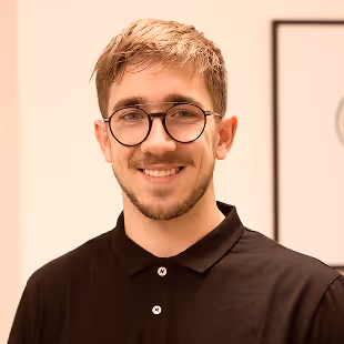 Smiling young man with short light brown hair and round glasses wearing a black collared shirt.
