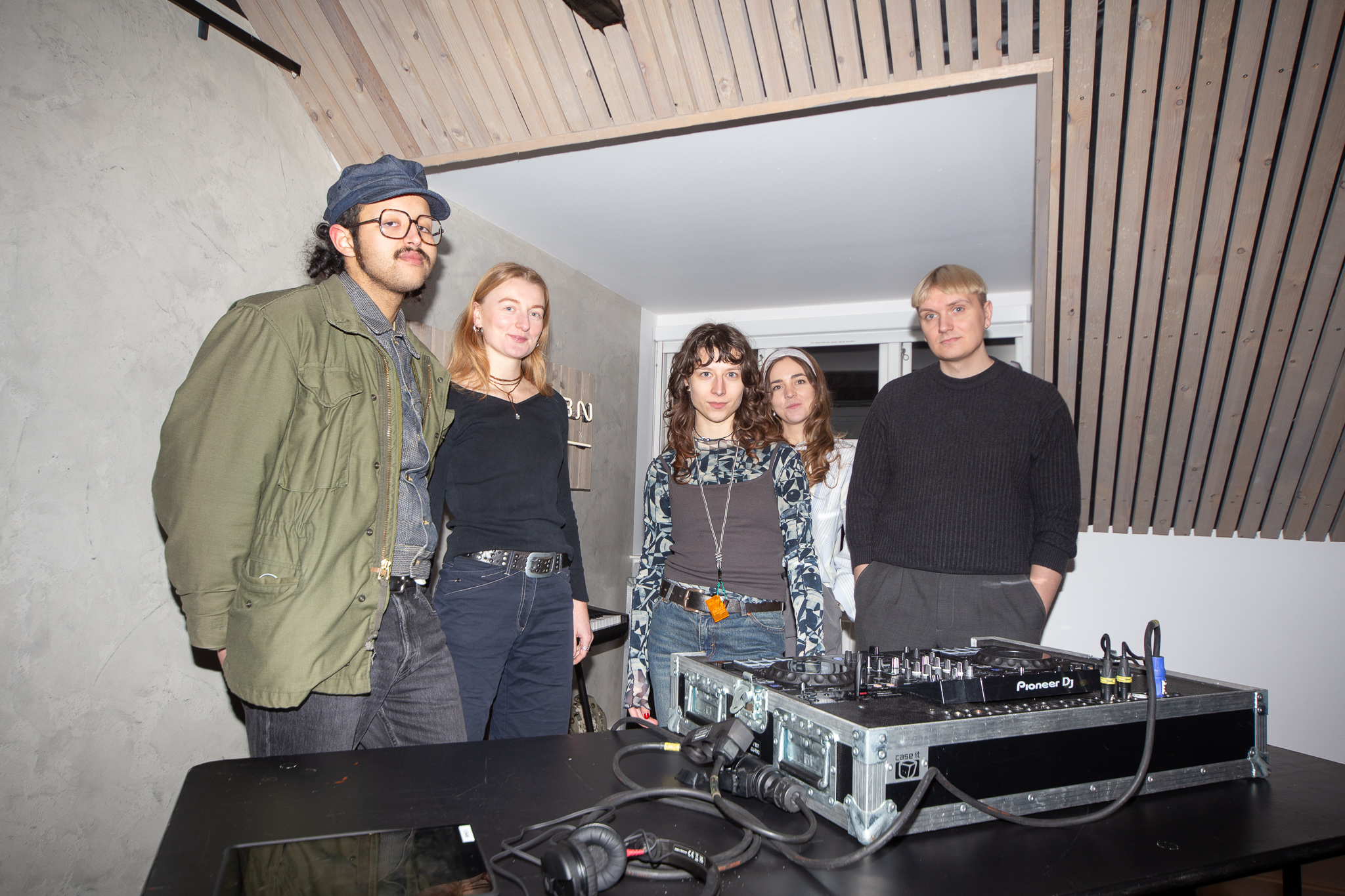 A group of five individuals standing together in a modern room with wooden ceiling details. They are dressed casually, with a DJ setup in front of them. The atmosphere appears relaxed and creative.