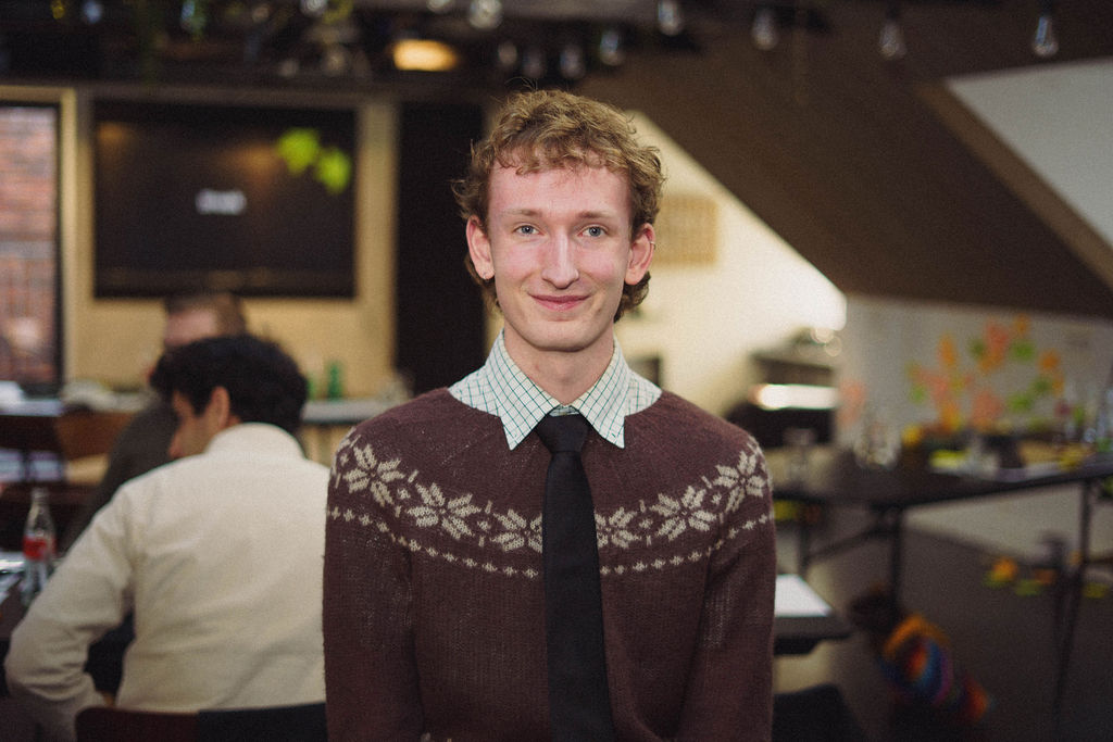 A young man wearing a brown sweater with a geometric pattern over a collared shirt and tie, standing in a social setting with people seated in the background. Dim lighting creates a casual atmosphere.
