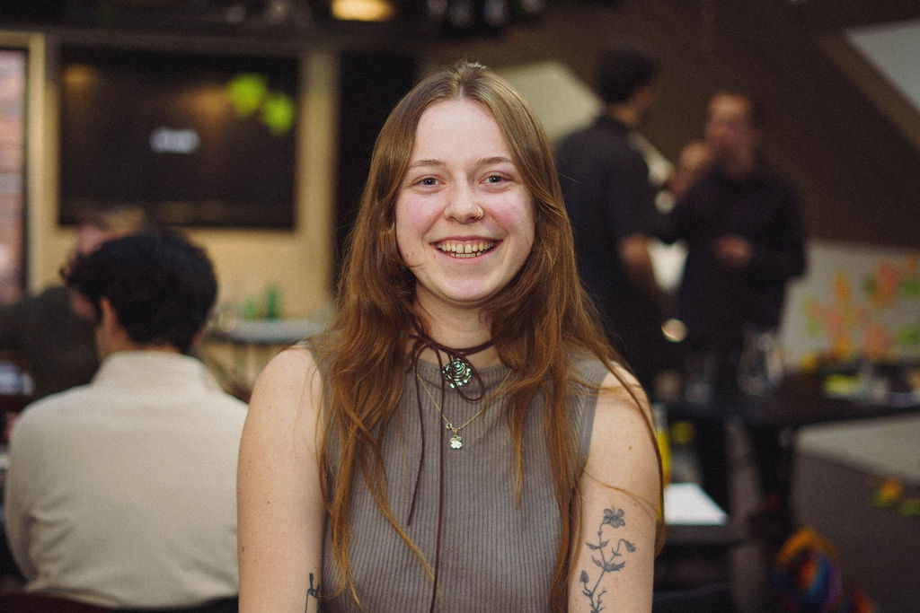 A young woman with long hair and a smile, wearing a sleeveless top, is seated in a café setting. In the background, other people are engaged in conversation. A screen is visible behind her.