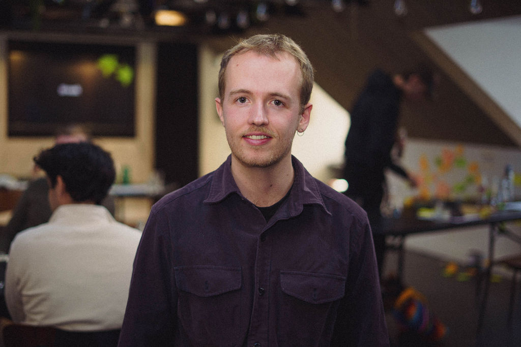 A young man with light brown hair standing in an indoor workspace, wearing a dark shirt. In the background, colleagues are engaged in discussions and activities. A screen is visible, showing a digital interface.