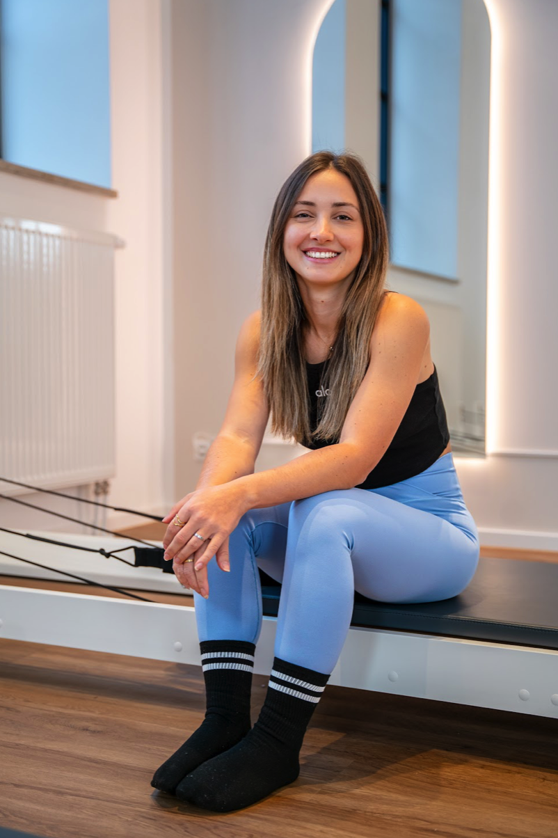 Junge sportliche Frau sitzt lächelnd in Sportbekleidung auf Reformer in einem ästhetischen Studio.