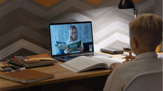 Child at a desk video calling a woman who is reading a book titled 'Family & Friends 3'.