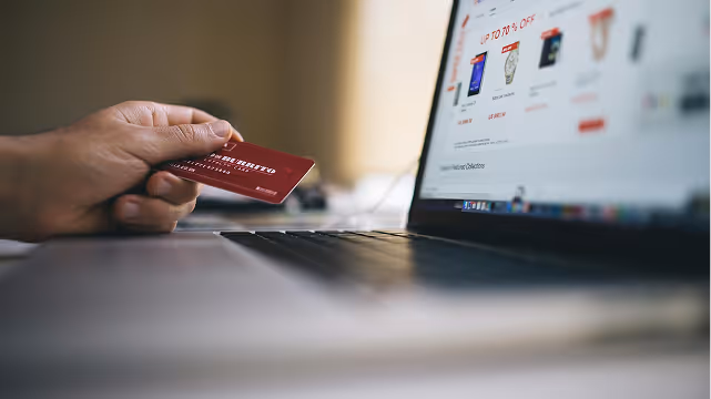 Person holding a red credit card near a laptop displaying an online shopping website with discount offers.