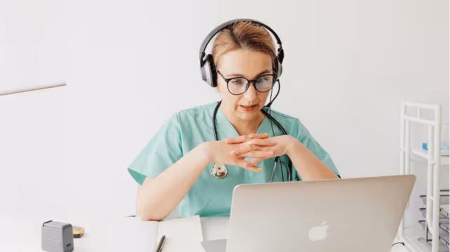Female healthcare professional wearing scrubs, headset, and glasses, sitting at a desk with a stethoscope around her neck, looking at a laptop.