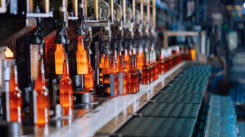 Conveyor belt with rows of amber glass bottles being processed in a bottle manufacturing factory.