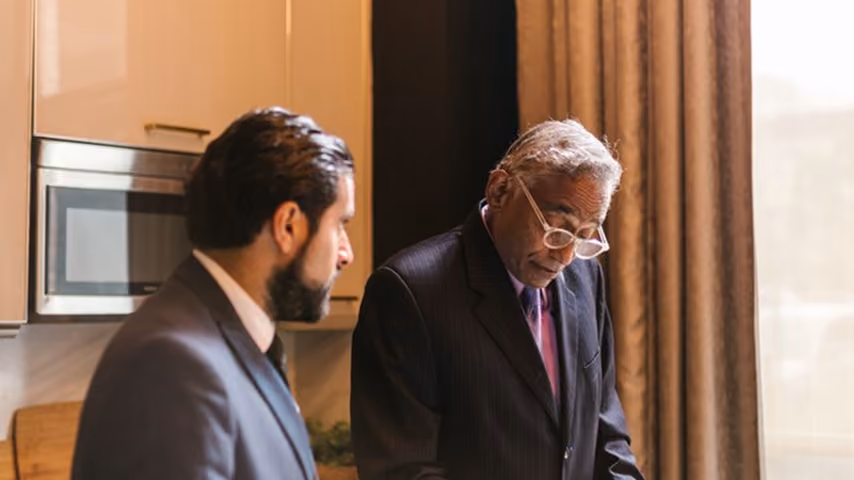 Two businessmen in suits discussing work documents in a modern office with a kitchen area.