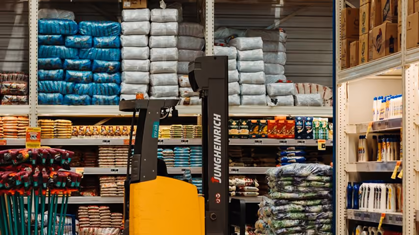 Yellow Jungheinrich forklift in warehouse aisle with stacked bags and stocked shelves.