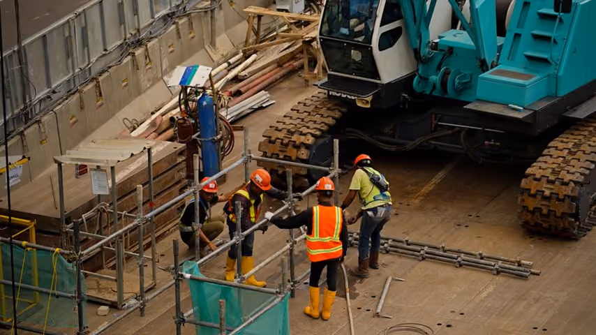 Four construction workers with helmets assembling scaffolding beside a large tracked excavator.