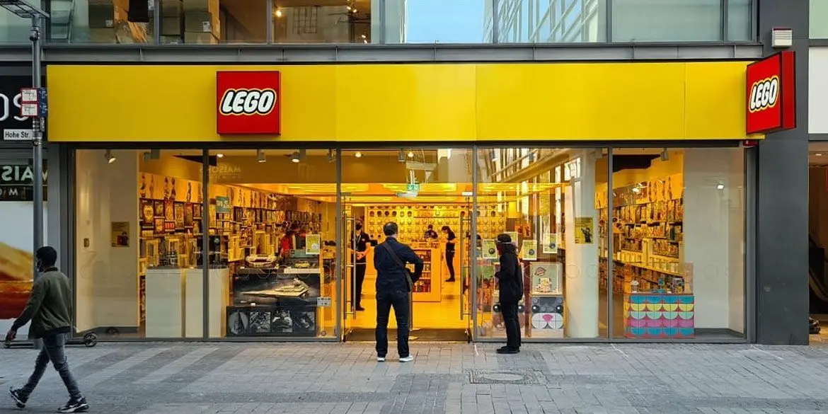 The exterior of a LEGO retail store with a bright yellow storefront sign and red LEGO logo, with shoppers visible through the glass entrance.