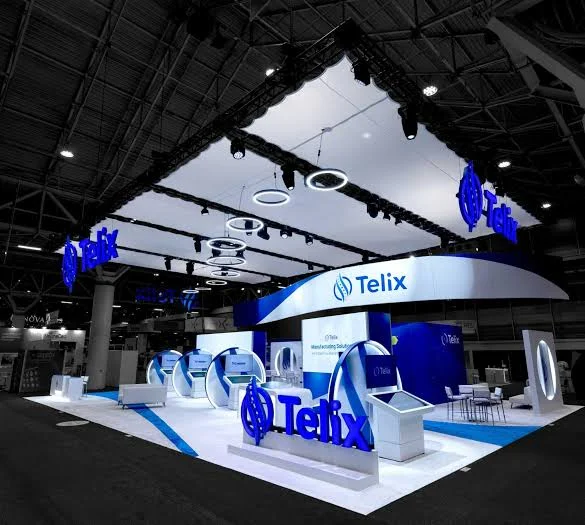 Telix island trade show booth with glowing blue logo letters, a white ceiling with ring lights, and touchscreen kiosks