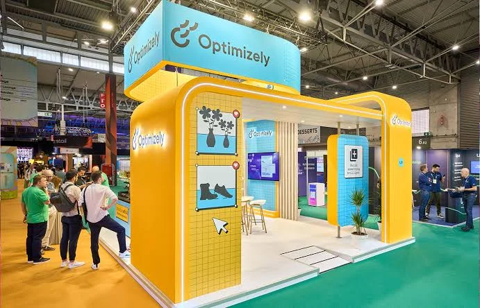 Optimizely trade show booth with a yellow arched frame, cyan header sign, and grid panels showing plant and boot icons