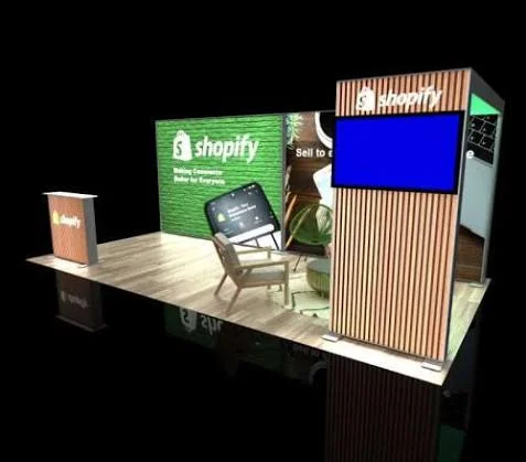 3D render of a Shopify trade show booth with a green brick wall, a wood-slat tower with a screen, and a lounge chair