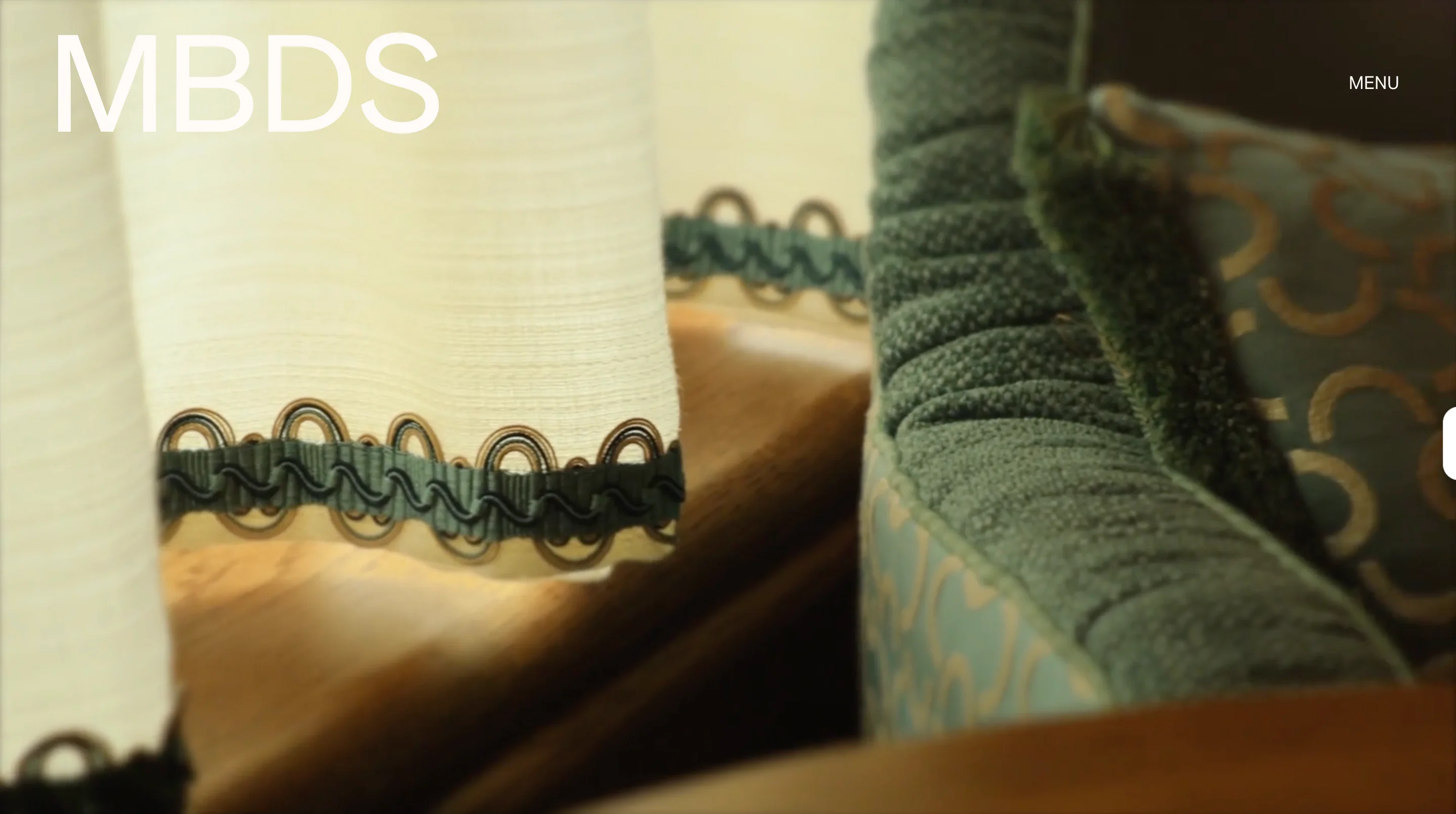 Martin Brudnizki Design Studio homepage with a close-up of cream curtain trim and a green velvet sofa behind the MBDS logo