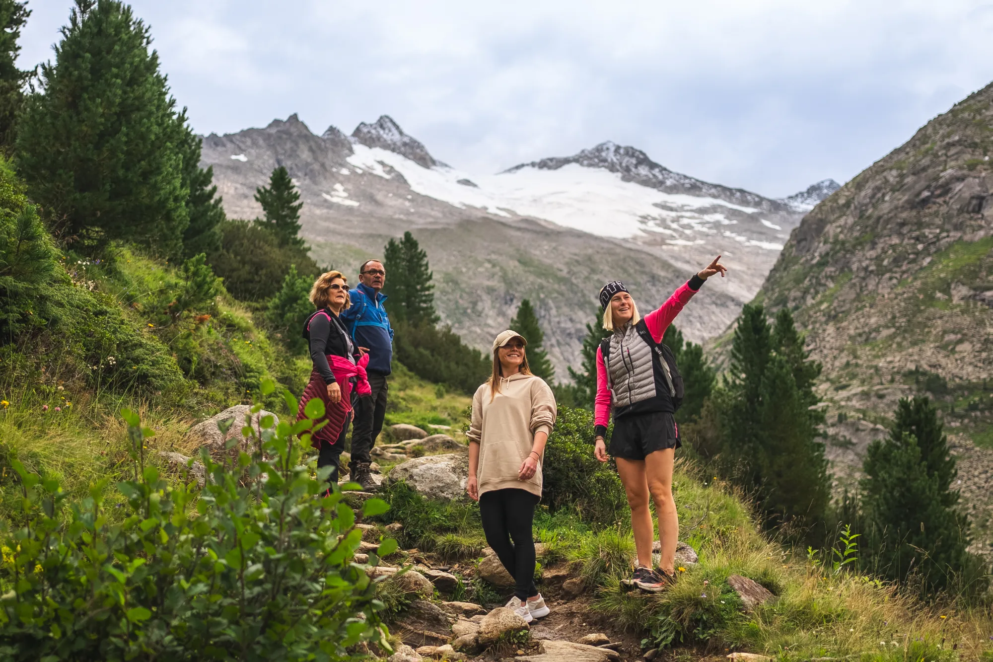 Vier Wanderer auf einem Steig zwischen Zirben, eine Frau zeigt auf die vergletscherten Gipfel im Hintergrund.
