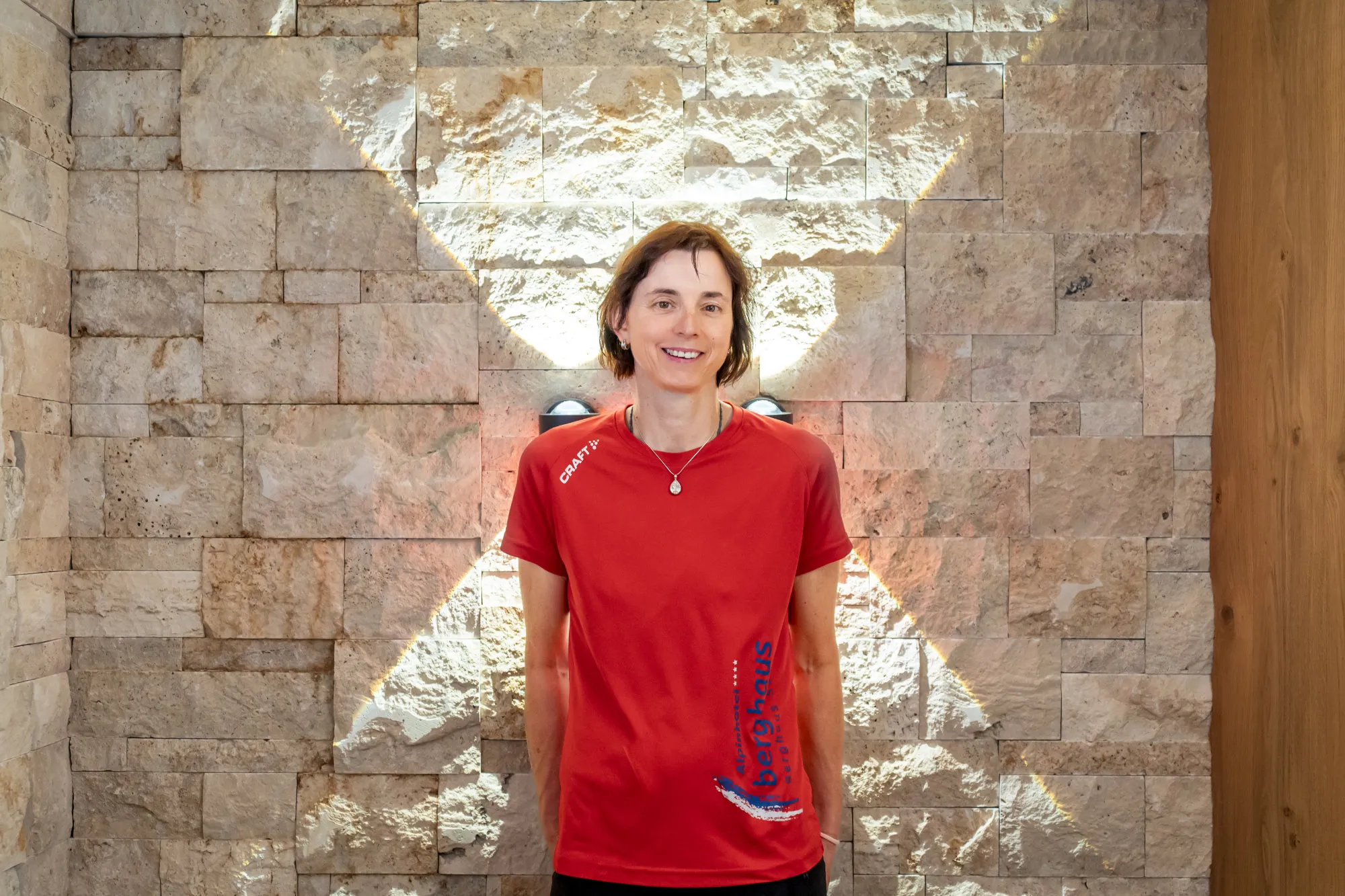 Frau in rotem T-Shirt lächelt vor einer Steinwand mit kreuzförmigem Lichtmuster.