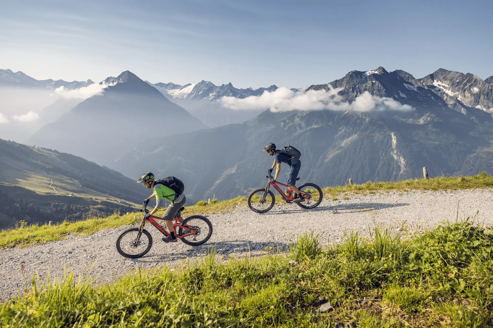 Zwei Mountainbiker auf einem Schotterweg am Berg, dahinter Gipfel und Wolken über dem Tal.