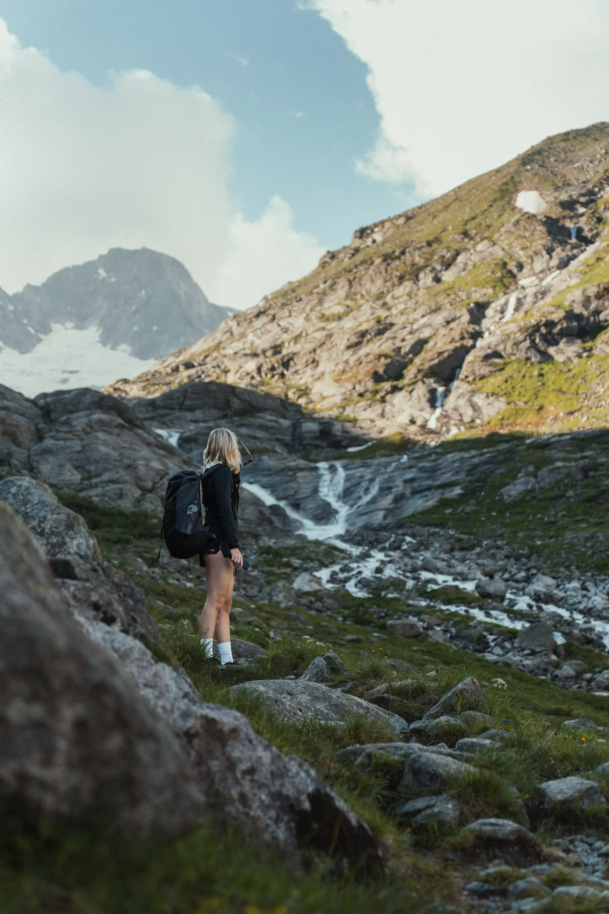 Wanderin mit Rucksack blickt über ein Hochtal mit Bachlauf und Schneefeldern.