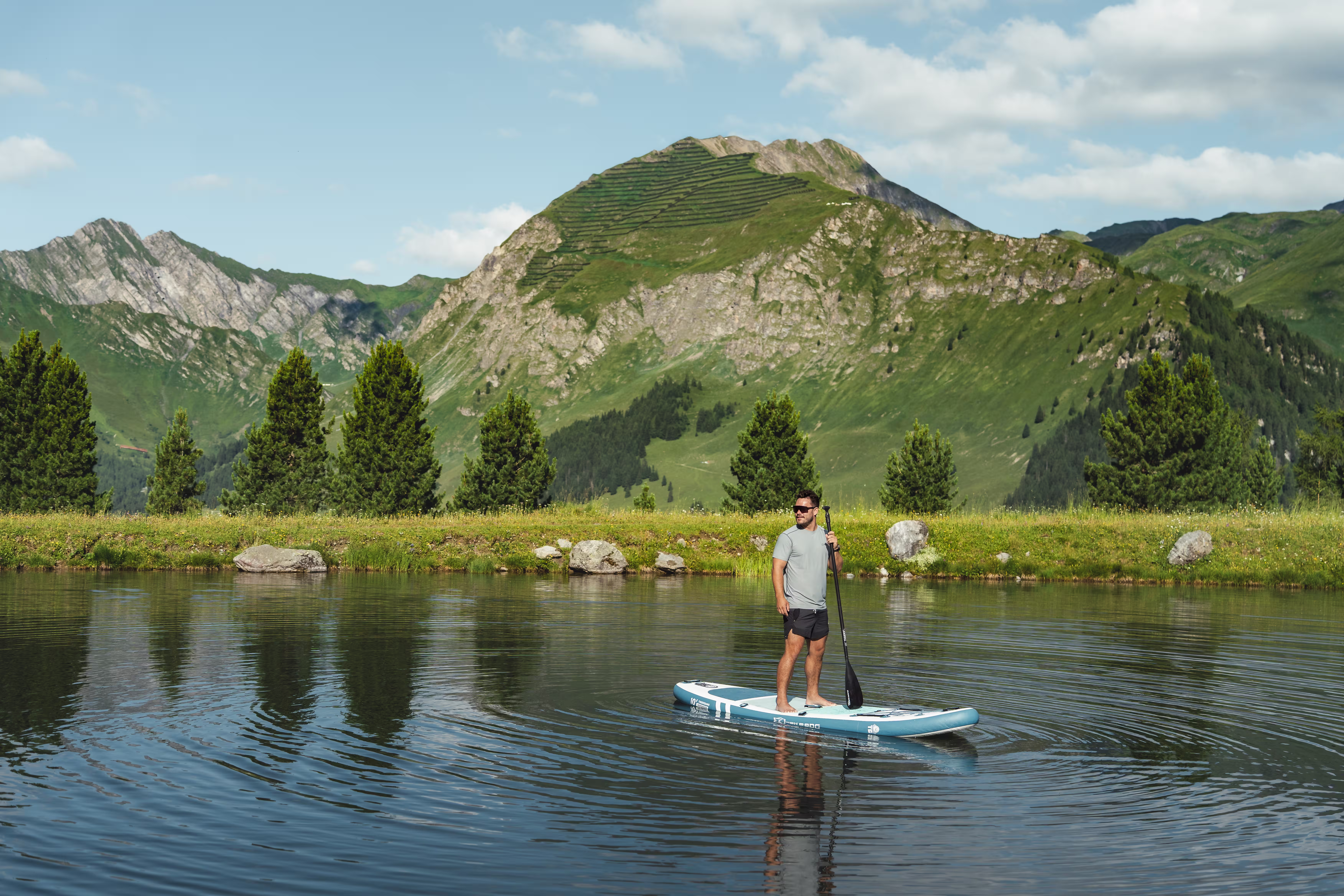 Mann auf einem Stand-up-Paddle-Board auf einem Bergsee, dahinter grüne Alphänge.